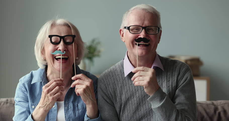 Cheerful elderly couple sit on couch hold funny birthday and party set, carnival accessories, fake glasses and mustaches on wooden sticks smile look at camera celebrate life event, fiesta feel happy Royalty-Free Stock Footage #1098773669