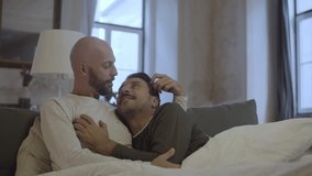 Happy homosexual men in pajamas hugging in bed in morning. Medium shot of smiling gay couple looking at each other with tenderness, talking and stroking each other. LGBT, love, romance concept - Powered by Shutterstock - Get 15% off with code: PIKWIZARD15
