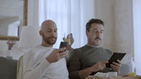 Happy gay couple having breakfast in bed and using gadgets. Cheerful homosexual men in pajamas reading news on tablet and phone, talking, spending morning together. LGBT, love, morning routine concept - Powered by Shutterstock - Get 15% off with code: PIKWIZARD15