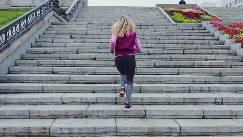 Fit girl in sportswear runs up urban stairs. Faceless sportswoman doing cardio workout on staircase outside. Blonde woman is in hurry, quickly overcoming distance along street stairway