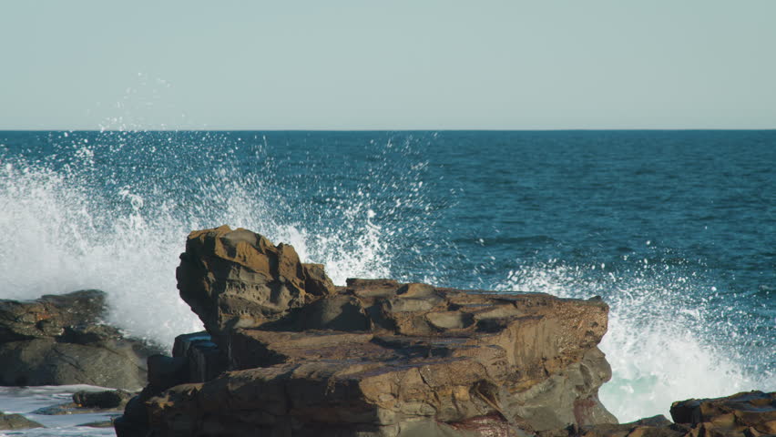 Large waves breaking against rocks with blue ocean in the background and clear sky on the horizon. Filmed at Phillip Island on the coast of Australia.
