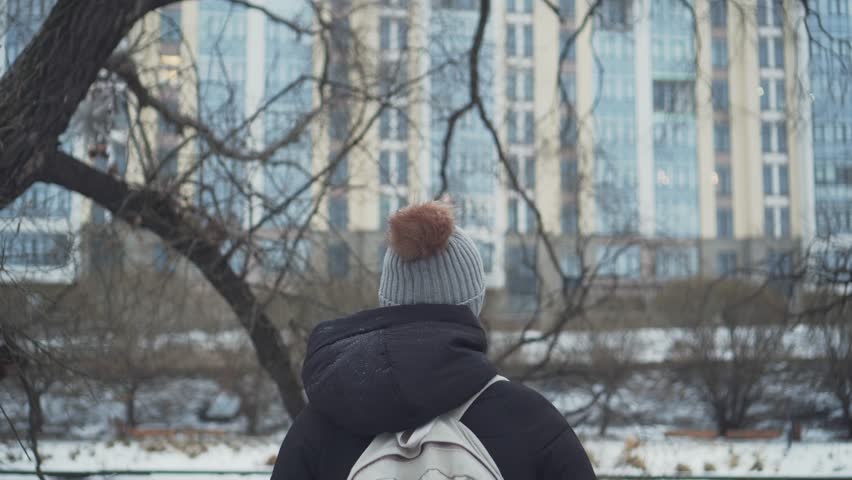 Rear view. A human in a hat on the background of an apartment building in snowy weather on the street. A woman is walking in a city park, it is snowing Royalty-Free Stock Footage #1098806375