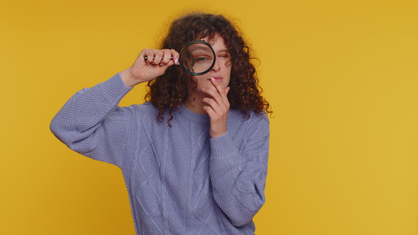 Investigator researcher scientist woman holding magnifying glass near face, looking into camera with big zoomed funny eyes, searching, analysing. Young curly haired girl on yellow studio background | Shutterstock HD Video #1098808551