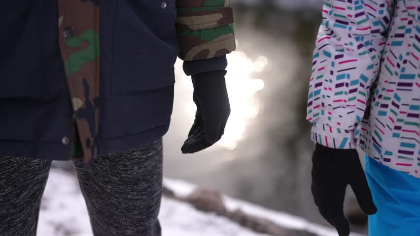 Unrecognizable teen couple holding hands at background of sunshine reflecting in river water. Happy loving boy and girl taking hands standing outdoors on cold winter day