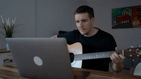 Man watches online lessons on laptop learning to play guitar. Male person with musical instrument sits at wooden table near gadget slow motion