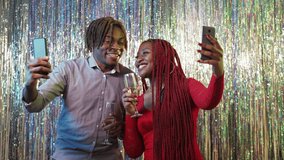 Online greeting. Excited couple. Festive celebration. Happy black man and woman taking video with champagne on mobile phones posing shimmering cascade curtain background.