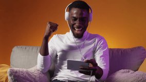 Video game online competition. Digital leisure. Excited guy in headphones winning losing playing on phone on couch in purple neon light on orange free space.