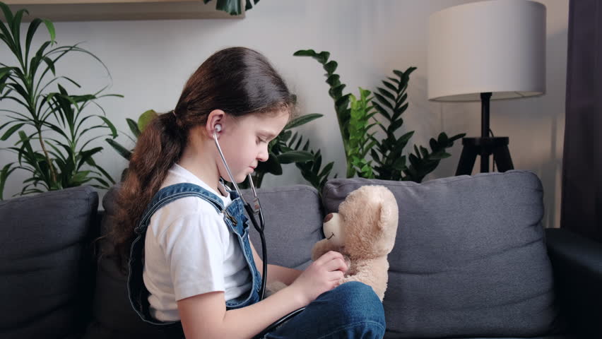 Little child plays with teddy bear sitting on cozy couch at home, childhood dream of becoming doctor, kid veterinarian examines toy doll with stethoscope, pretend heartbeat, daughter treats friend Royalty-Free Stock Footage #1098814487