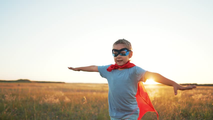 Happy child in summer in park at sunset. Girl in red superhero costume plays in meadow in grass. Active lifestyle in nature in spring. Child plays superhero at picnic in park. Childhood dream concept | Shutterstock HD Video #1098814663