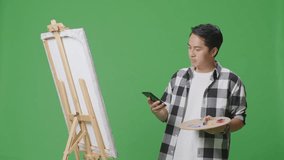 Asian Male Artist Having A Video Call On Smartphone While Painting On Canvas By Oil Paints And Brush In The Green Screen Studio
