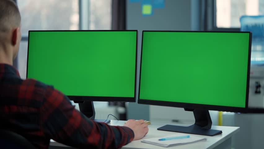 Young Man Working On Computer With Two Green Screen Mock Ups Sitting At Desk In Office