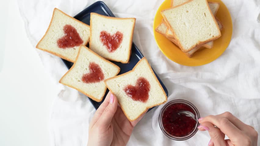 Slice of bread with strawberry jam in heart shape in the girl hand. Woman cooking a sweet breakfast - tasty sandwiches for Valentines day. Female hands apply heart shaped red confiture to white toast Royalty-Free Stock Footage #1098822355