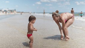 Mom plays and splashes on her baby who is laughing while standing in the sea under the summer sun on vacation