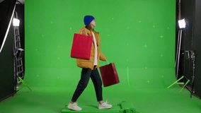 Young caucasian woman in warm clothes walking with shopping bags on a Green Screen, Sale day. Woman buyer on Chroma Key. 4k UHD side view video