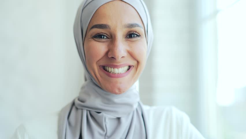 Closeup portrait of a pretty young Muslim woman in hijab standing and smiling indoors at home Headshot of an Islamic happy female girl looking at camera on a light room background close up | Shutterstock HD Video #1098825447