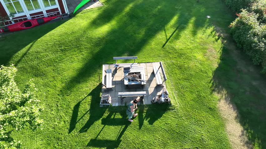 Women in green summer dress brushing cut grass from barbeque place after grass trimming, prepare barbeque with four benches and square fireplace for party. Aerial video