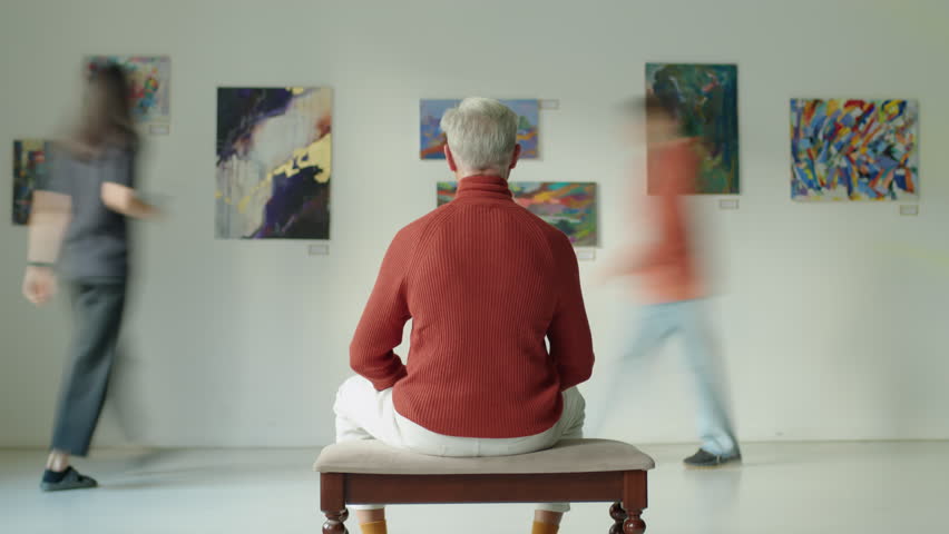 Time lapse of man sitting on bench in exhibition hall and looking at paintings while visitors walking around art gallery
