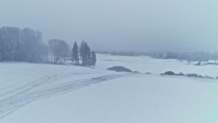 Aerial drone shot over icy road and farmlands covered with snow while heavy snowing on a foggy winter day.