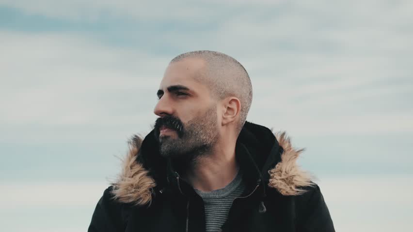 A handsome man stands on a mountain, looking at the breathtaking landscape of clouds on the horizon. Captured in slow motion, this close-up shot showcases his thoughtful expression and rugged style.