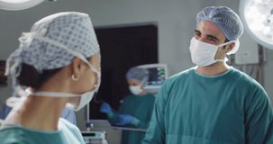 Video of asian female surgeon in face mask and diverse colleague talking and smiling in theatre. Hospital, medical and healthcare services.