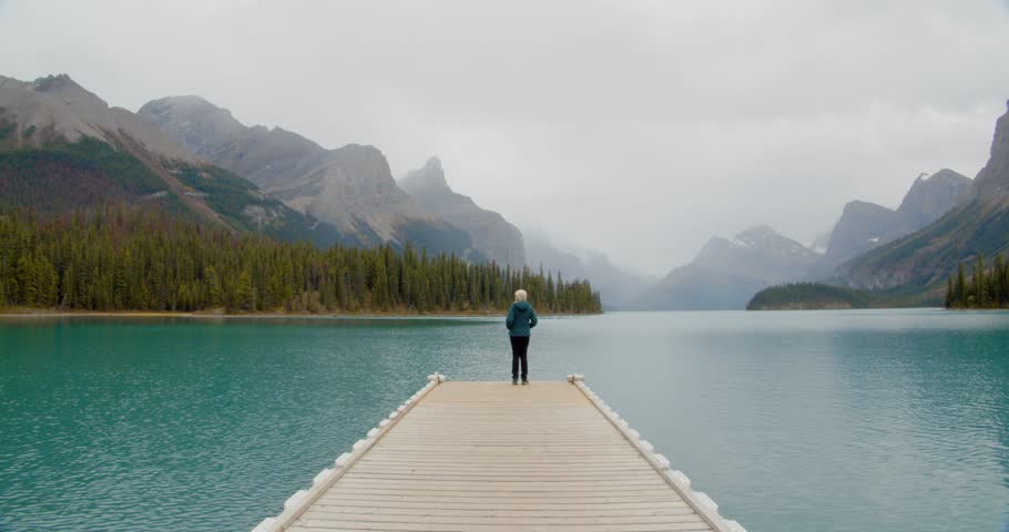 Outdoorsy woman looking mountain the horizon on the edge of a small pier floating on a stunning green water glacial lake nestled in the Canadian rocky mountains