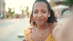 Closeup portrait of smiling young woman with ice cream in her hands making a video call on mobile phone on urban city background. Close-up of happy girl using a smartphone to communicate. Backlight