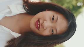 VERTICAL VIDEO: Close-up young Asian smiling woman with long brown hair wearing white t-shirt posing for the camera in the park . Girl touches her hair and smiles at the camera