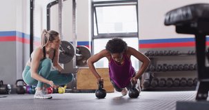 Video of diverse female fitness trainer instructing woman doing push ups on kettlebells at gym. Exercise, fitness and healthy lifestyle.