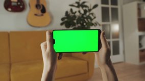 Holding Phone in Horizontal Positiion. Watching Content, Videos, Blogs. Handheld Camera: Point of View of Woman at Modern Room Using Phone With Green Mock-up Screen. Chroma Key Surfing Internet.
