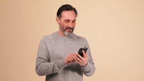 Happy adult bearded man in grey sweater talking by video call on mobile in the beige studio