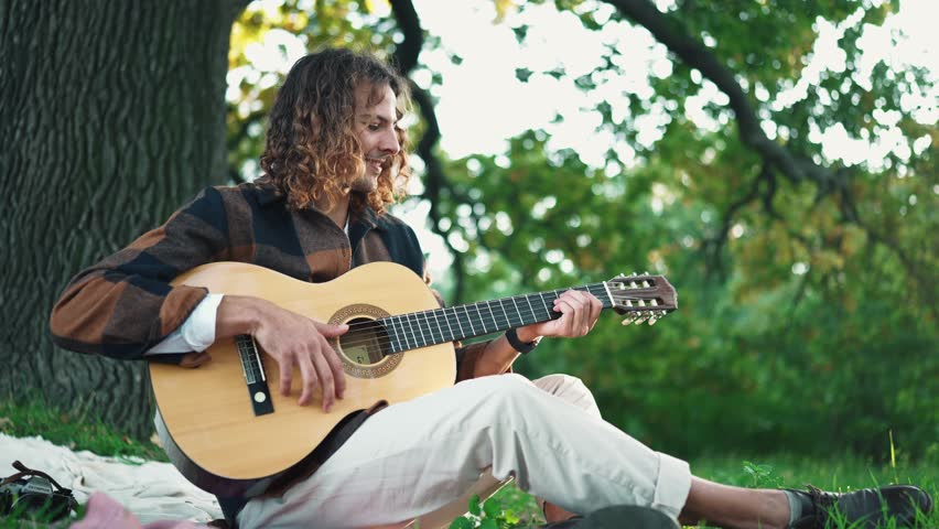 Funny curly haired man playing on guitar and singing on blanket at a picnic in the park