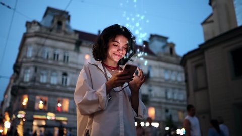 Стоковое видео: Beautiful Smiling Woman Using Smartphone on a City Street at Night. Wireless communication network concept. Mobile technology. Visualization of Information Lines Flying from Mobile Phone