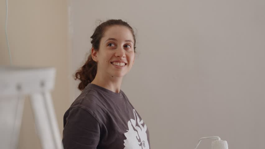 Woman smiling and holding paint roller repainting a room in her new or old house. Cheerful young female doing remodeling and painting walls in her apartment or home. Looking around at the work ahead Royalty-Free Stock Footage #1098851547
