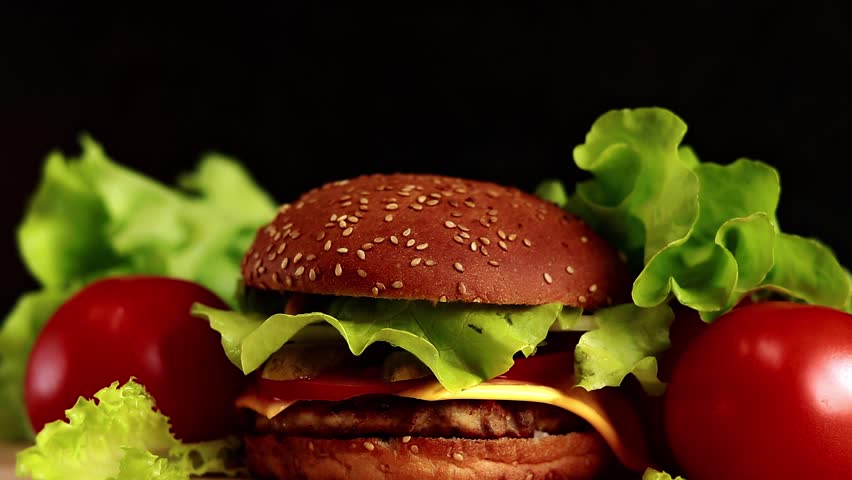 Burger with cutlet, melted cheese, salad and vegetables on a dark background. Burger and fresh vegetables: lettuce and tomatoes. Fast food. Smooth video from bottom to top | Shutterstock HD Video #1098858549
