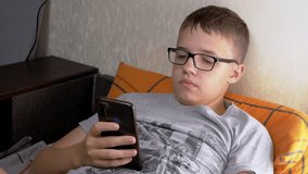 Child in Glasses Lying on a Sofa, Playing Video Games on a Smartphone in Room. Tired cute boy with poor eyesight looks at mobile phone screen before going to sleep. Internet, reading, working online.