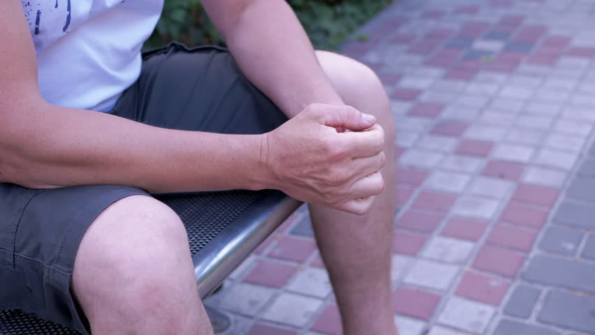 Male Rubbing Hands Sitting on a Bench in the Urban Park. A moment of waiting, excitement, anxiety, and emotional stress, trouble, nerves, and dread. Massage of hands, joints, fingers, skin. Outdoors.