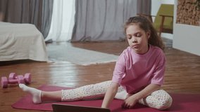 Funny adorable little girl with dark curly hair wearing pink clothing doing sport alone at home in living room in front of open laptop. Home gymnastic. Kids sport. Online training.
