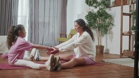 Young Hispanic Woman teach doing stretching exercises training her little cute daughter, sit floor in cozy living room perform in front of open laptop. Happy activity in family concept.