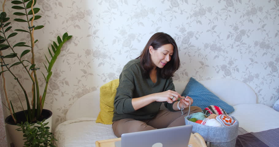 Close-up video of woman crocheting something small. Anti-stress hobby. Tranquil leisure at home. Handicraft. Online lessons for amateurs on laptop.