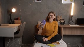 A girl student sits in a college dormitory on a bed in an orange sweater with glasses looking to make a video call online using a smartphone.