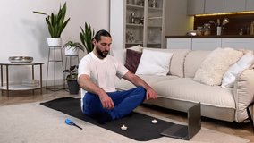 Tranquil mid adult bearded man meditating, following instruction online yoga classes via laptop at home. Peaceful sporty male breathing deeply, siting with closed eyes on exercise mat in living room