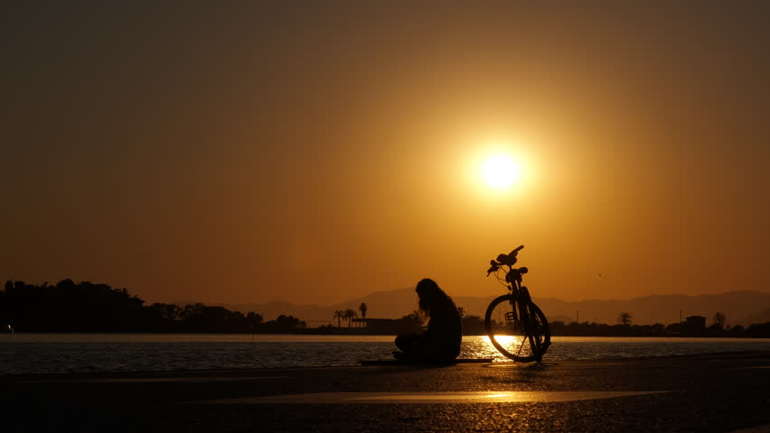 Meet the sunset with a bike by the bay. Silhouette of a girl in a lotus position on the embankment