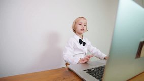 Cute blonde boy in stylish black tie bow and bright trousers looks at the laptop screen and talks to a teacher repeating words and sounds. Online speech lesson at home for a 5 years old boy
