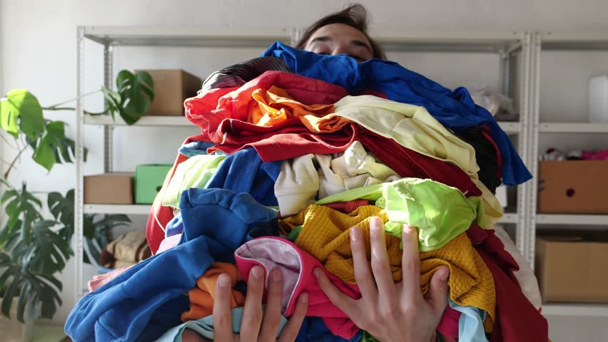 Fast Fashion and Textile Waste. A large pile of old clothes in the hands of a young man. Waste is at the heart of fashion both the physical act of discarding materials and clothes Royalty-Free Stock Footage #1098887499