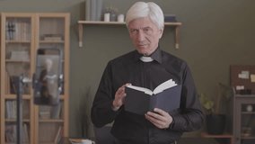 Senior priest in black shirt and white clerical collar preaching sermon at home and recording video of it on smartphone standing on tripod