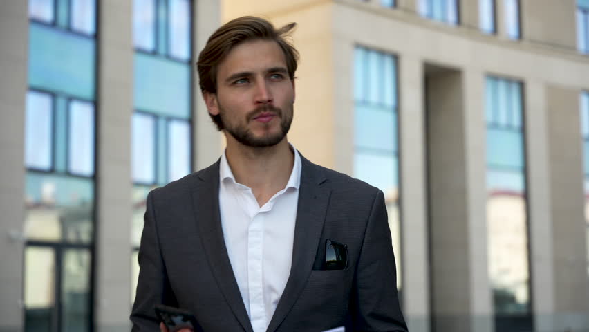 Portrait of successful businessman wearing stylish suit holding documents walking outdoor near the office buildings. Man holding smartphone typing message. Business concept | Shutterstock HD Video #1098889099