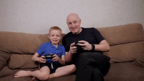 Cheerful father and son play video games on consoles at home on the sofa in the living room.