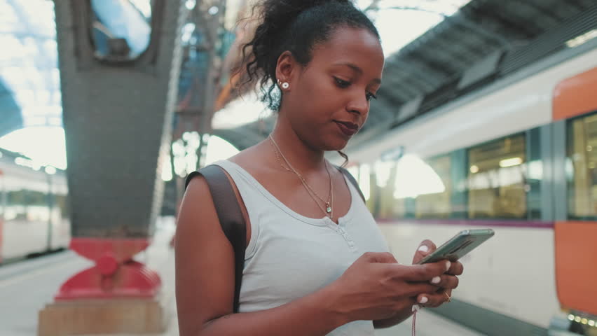 Young woman stands at the railway station on the background of trains, uses smartphone
