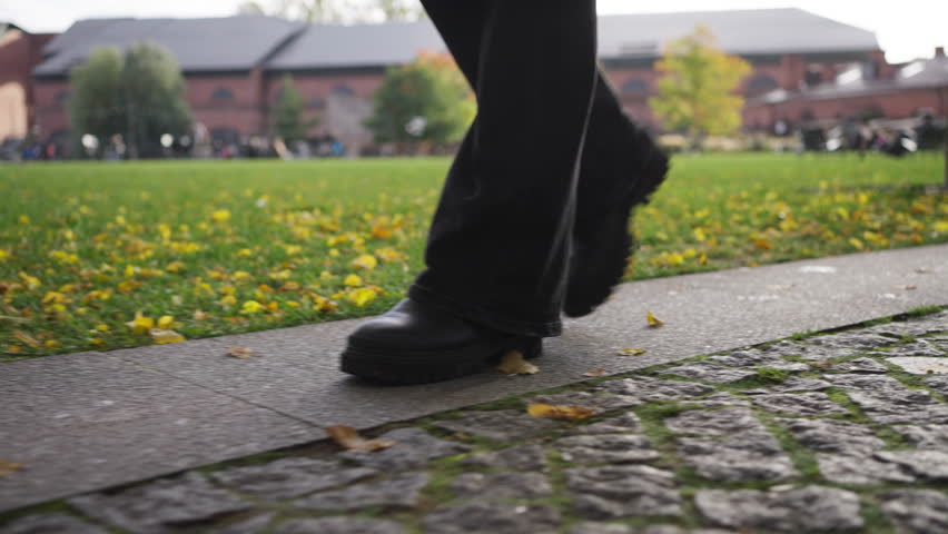 Close-up female legs in black boots and jeans walk along the path against the background of a green lawn with autumn leaves on a sunny day