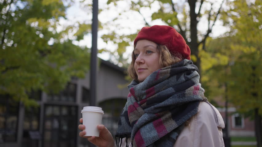 Woman in a red beret walks through a city park against the backdrop of yellow trees and enjoys drinking coffee from a paper cup in slow motion | Shutterstock HD Video #1098894503